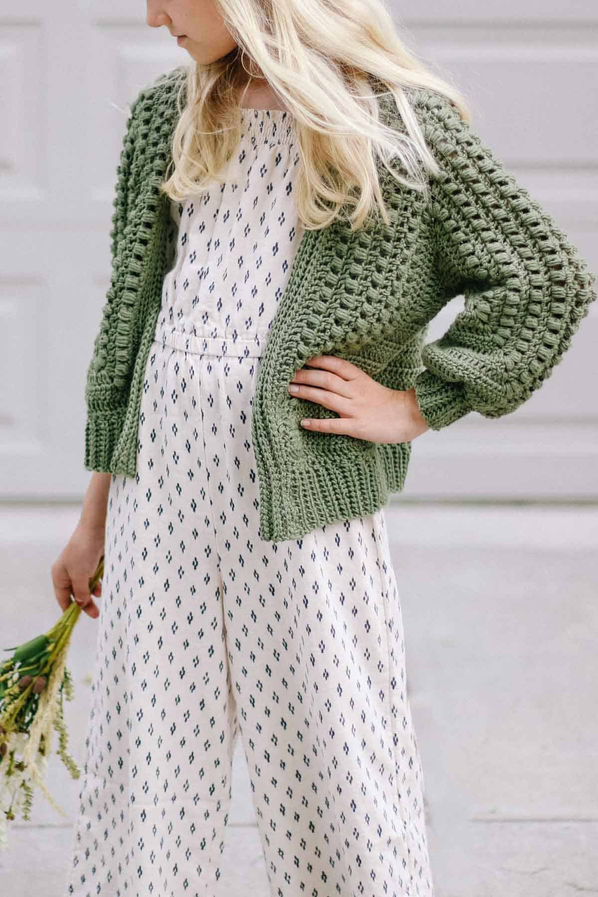 Blonde girl wearing an olive green crochet cardigan and holding a bouquet of flowers.