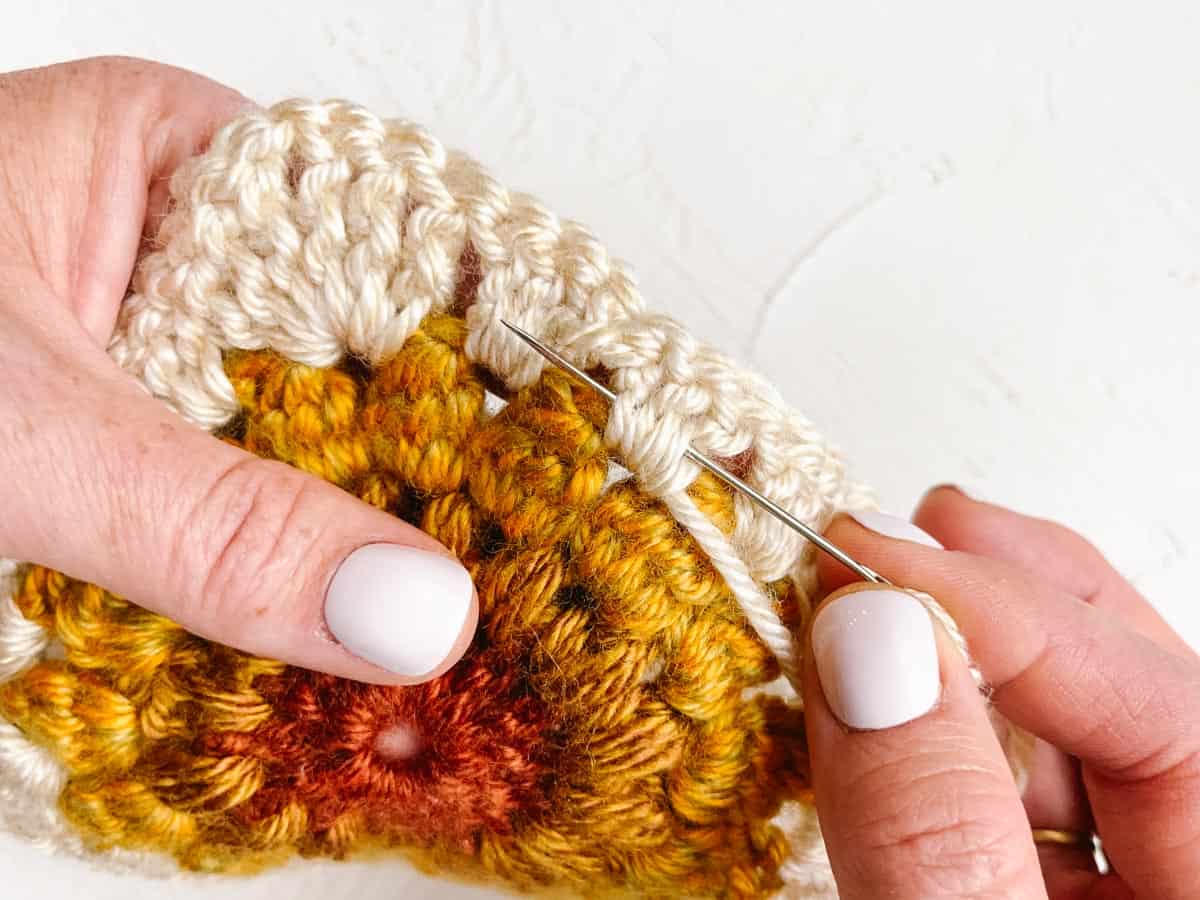 Tapestry needle weaving yarn end through crocheted granny square corner.