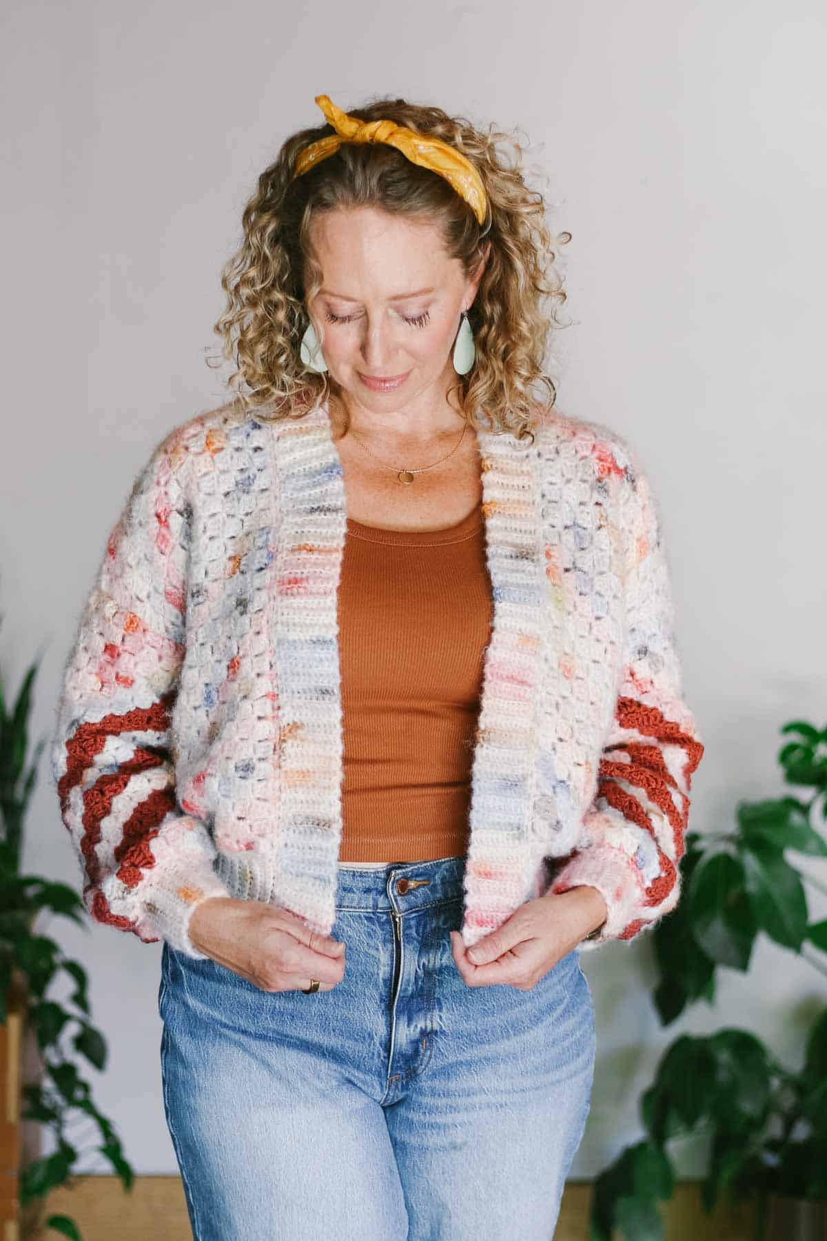 A colorful c2c crochet cardigan worn by a woman paired with a brown top and denim jeans with her hands on the front ribbing of the cardigan.