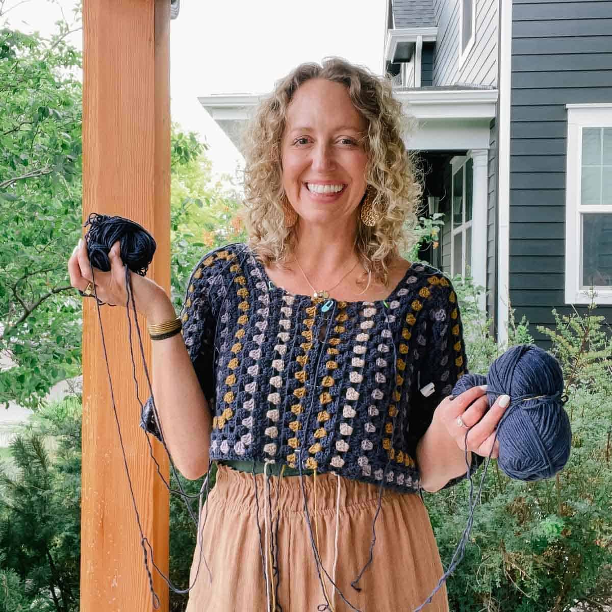 An in-progress crochet hexagon dress worn by a blonde woman while holding yarn skeins.