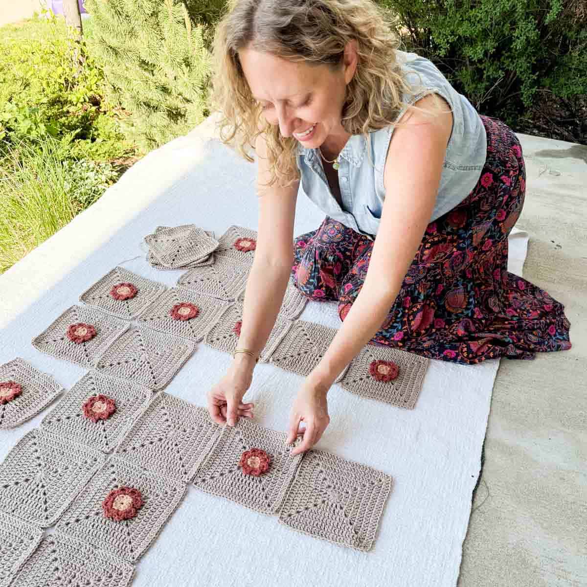 A blonde woman arranging crochet square motifs and flower motifs laid on the ground to create a flower cardigan.