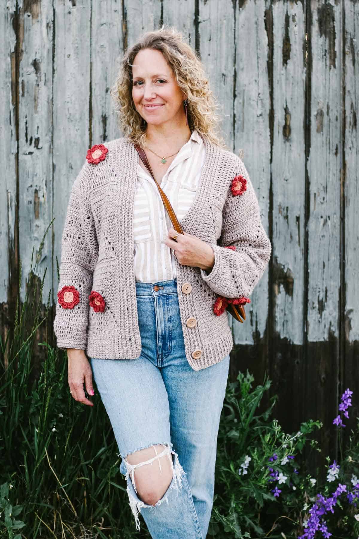 A crochet flower cardigan worn by a blonde woman paired with a stripes button down, ripped denim jeans and a brown leather sling bag standing on a garden.