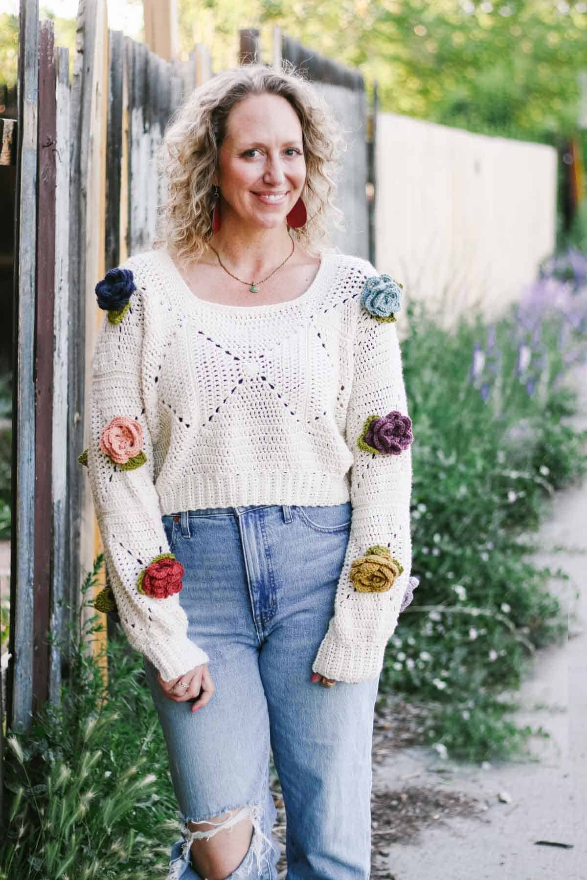 A crochet flower pullover worn by a blonde woman paired with ripped denim jeans standing on a garden.

