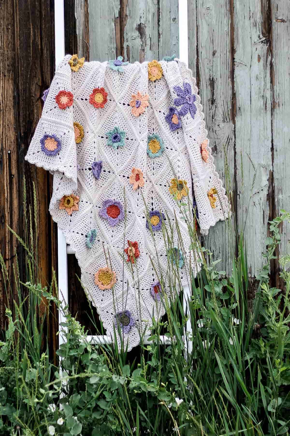 A crochet flower blanket draped on a stand by the wooden fence.