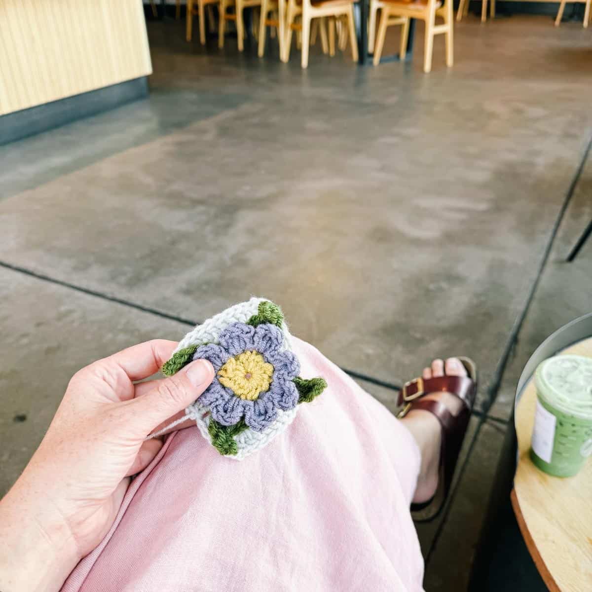 An in-progress crochet flower motif with a square held by a woman sitting on a cafe.