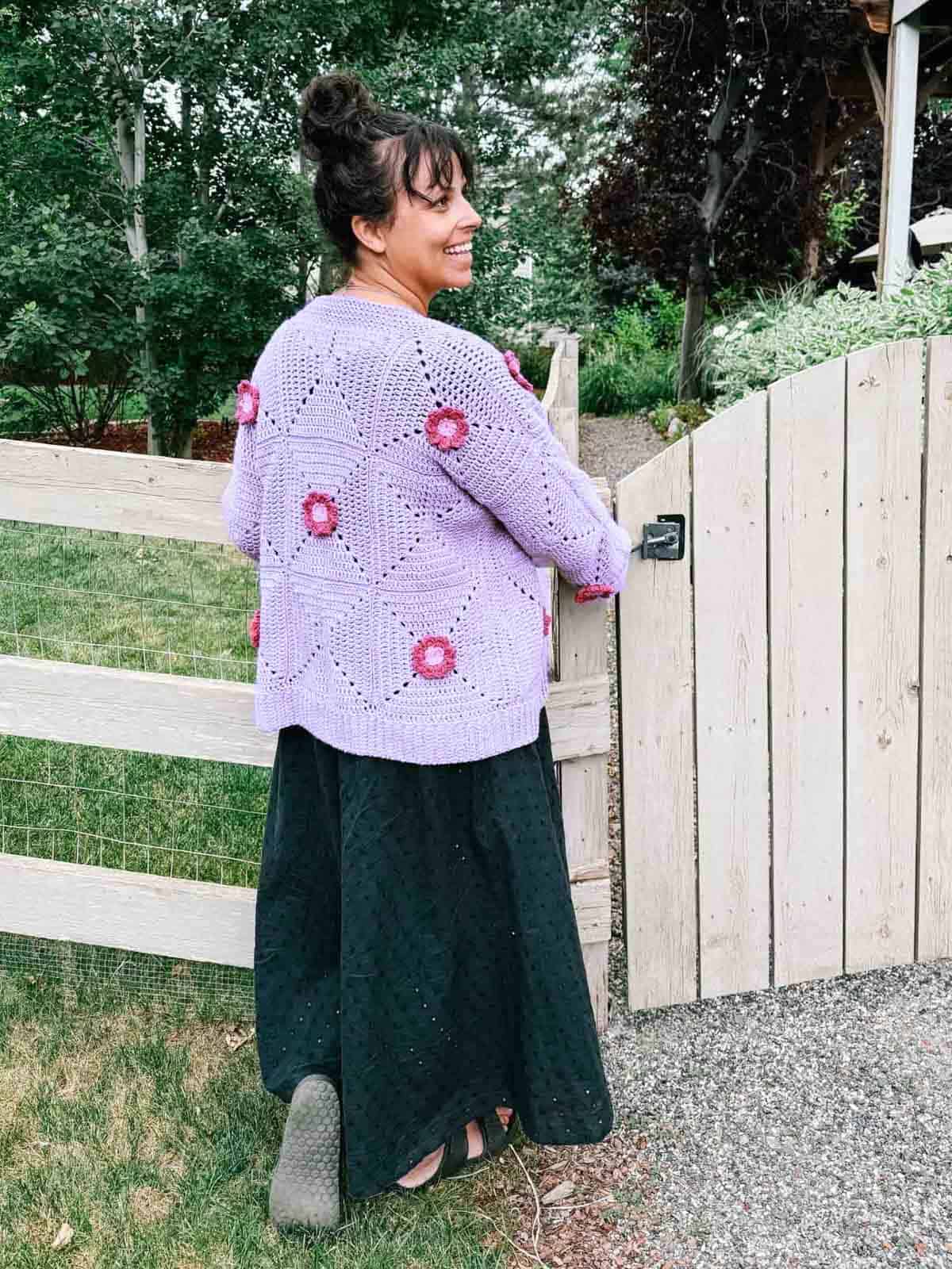 A lilac colored crochet flower cardigan worn by a woman paired with a long, flowy skirt standing on a garden.
