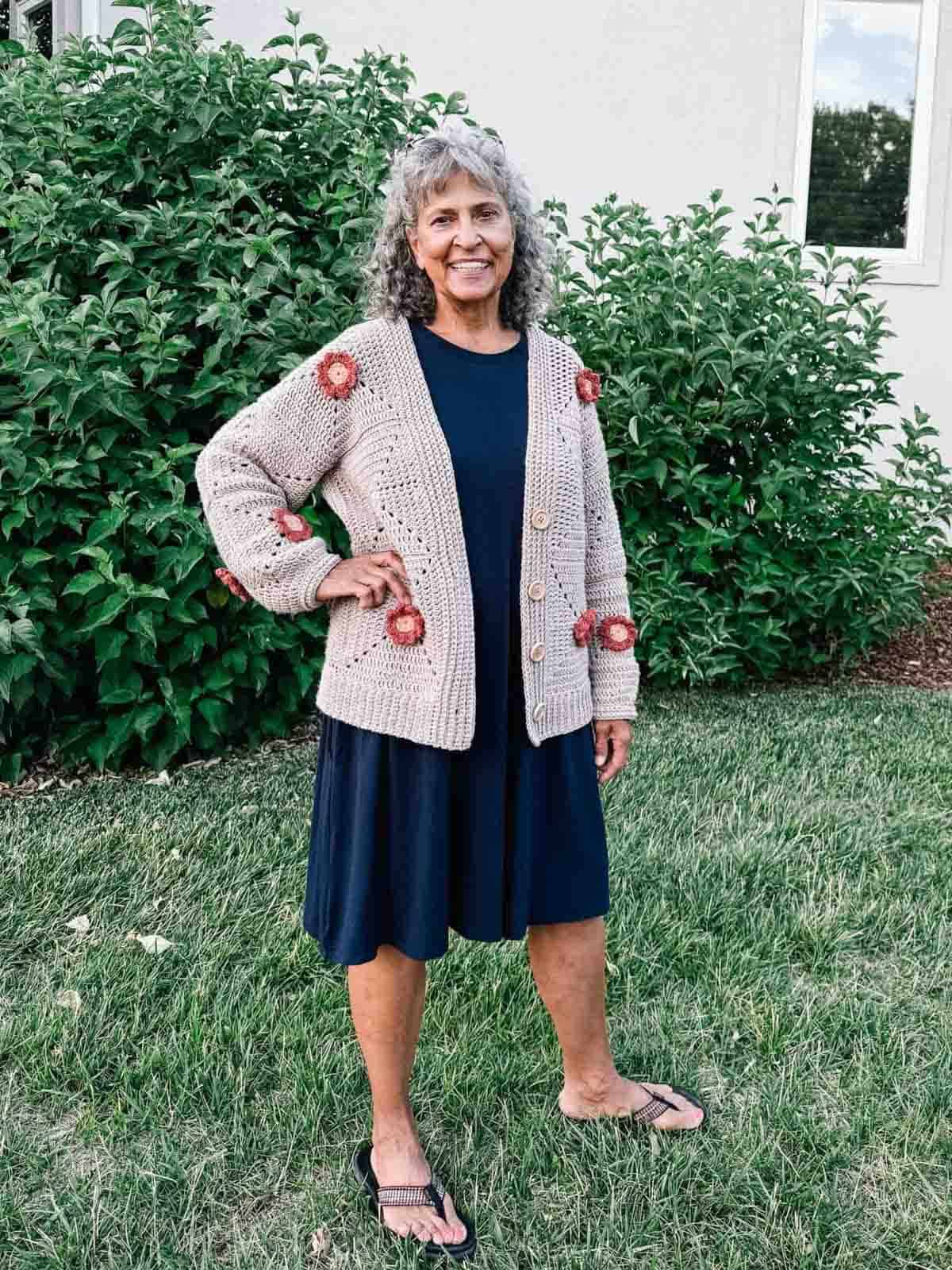 A crochet flower cardigan worn by a woman paired with a navy dress and a pair of slippers while standing on a garden.
