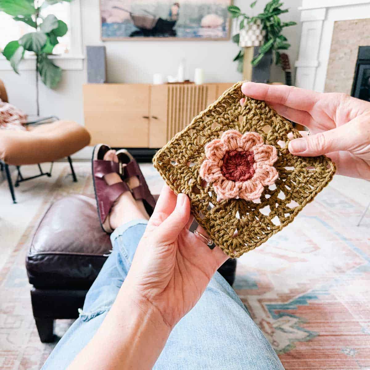 An in-progress crochet flower motif with a square held by a woman sitting on a living room.
