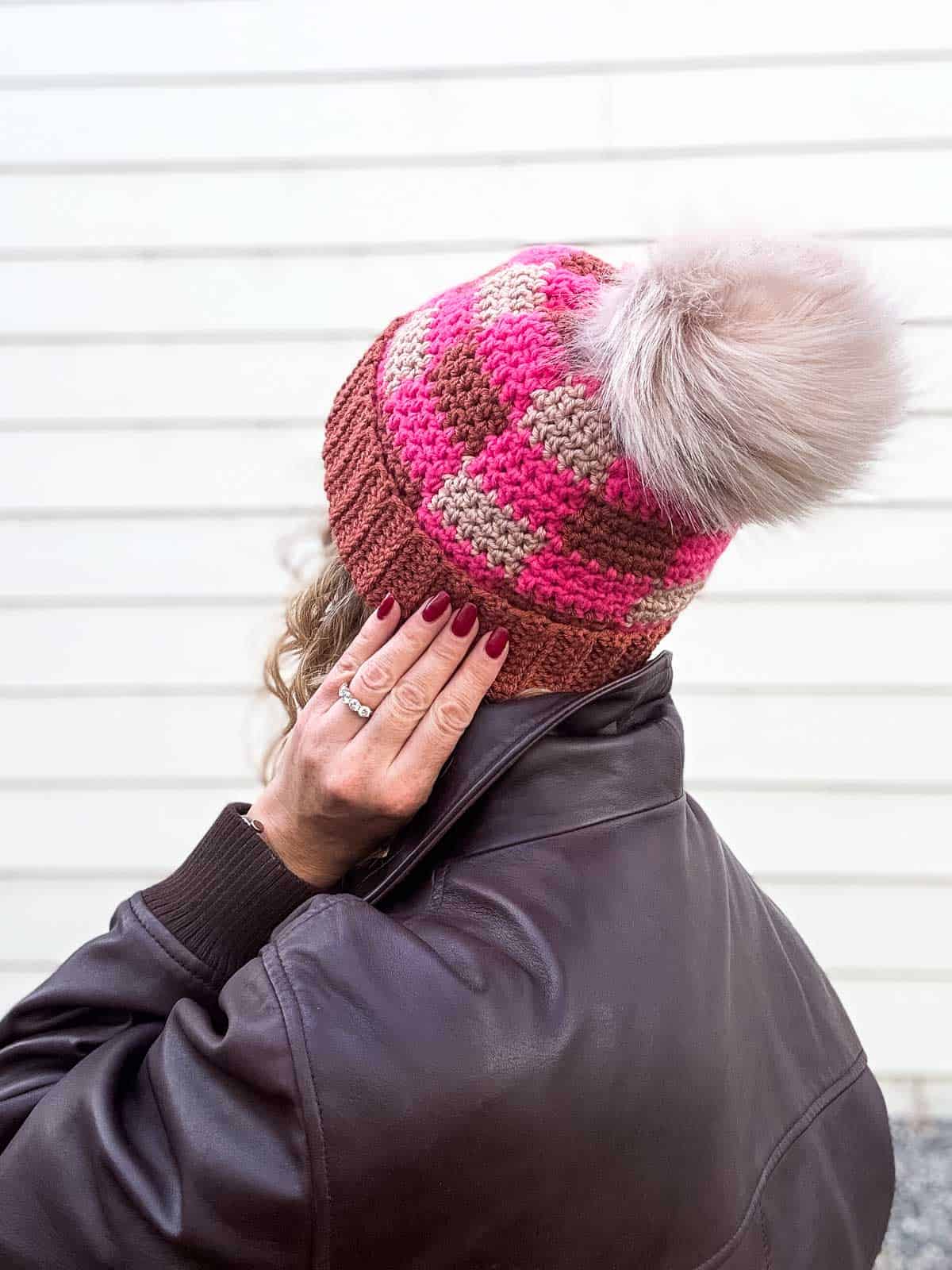 Back of woman wearing a pink plaid crochet beanie with a fur pom pom.